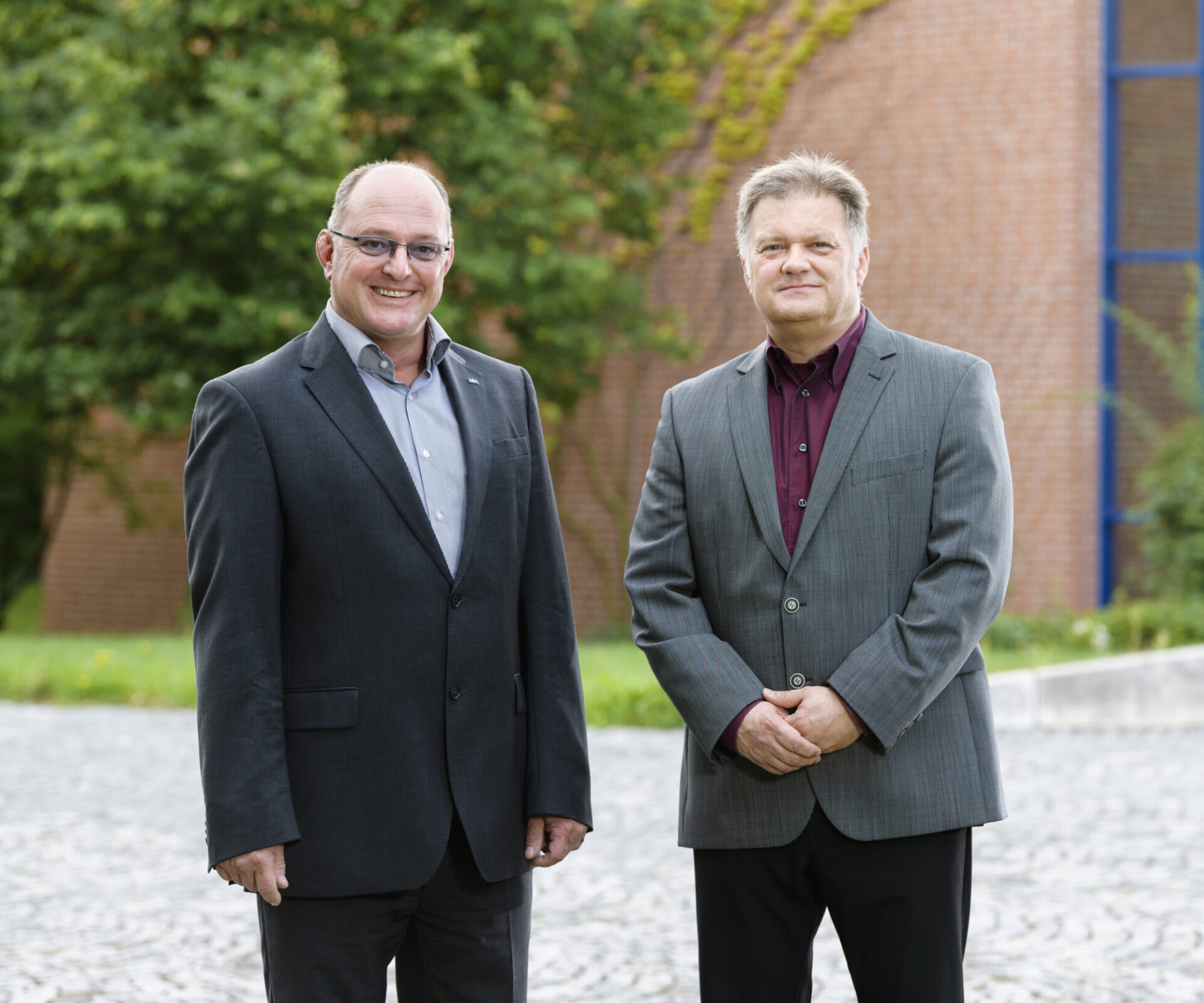 Das Bild zeigt zwei Personen im Freien auf einem gepflasterten Untergrund, im Hintergrund sind Grünflächen und ein Backsteingebäude zu sehen. Die linke Person trägt einen dunklen Anzug mit hellblauem Hemd, die rechte einen grauen Anzug mit bordeauxfarbenem Hemd.