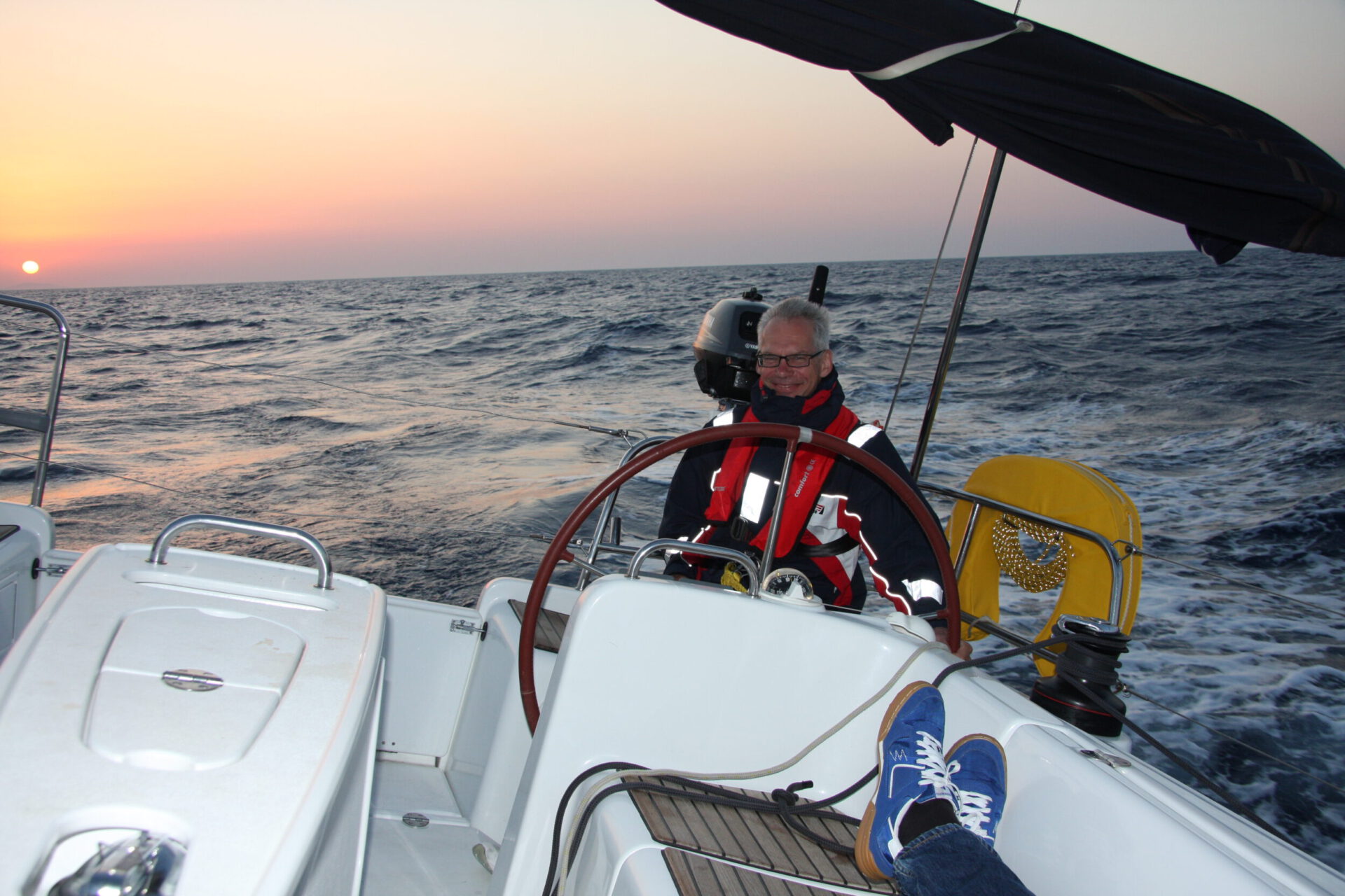 Das Bild zeigt eine Person am Steuer eines Segelboots, die das Boot über das Meer lenkt. Sie trägt eine rot-schwarze Jacke mit reflektierenden Streifen sowie eine Schwimmweste. Am Horizont geht die Sonne unter und taucht Himmel und Wasser in orange-rosa Licht.