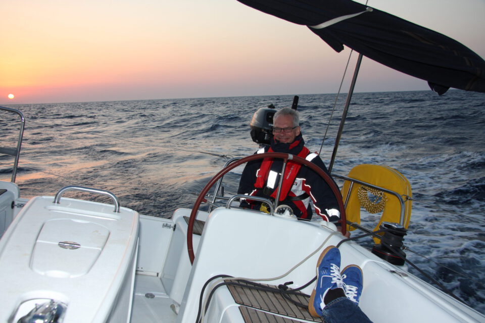 Das Bild zeigt eine Person am Steuer eines Segelboots, die das Boot über das Meer lenkt. Sie trägt eine rot-schwarze Jacke mit reflektierenden Streifen sowie eine Schwimmweste. Am Horizont geht die Sonne unter und taucht Himmel und Wasser in orange-rosa Licht.