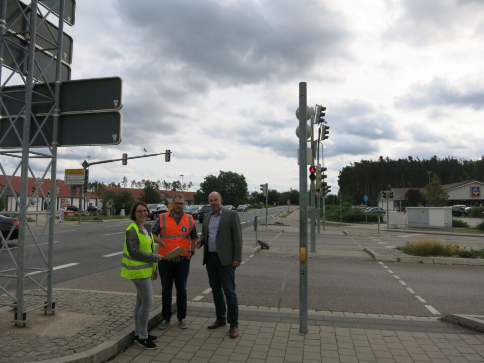 Das Bild zeigt drei Personen auf einem Gehweg in der Nähe einer Ampel und von Verkehrsschildern. Zwei tragen Warnwesten, eine gelb und eine orange, die dritte Person ist in einem Sakko gekleidet. Im Hintergrund sind eine Straßenkreuzung mit Ampeln, Schildern, Gebäuden und geparkten Autos zu sehen, der Himmel ist bewölkt.