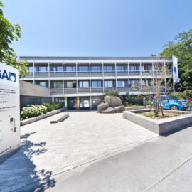 Das Bild zeigt ein modernes Bürogebäude mit drei Etagen und großen Fenstern, umgeben von Bäumen und Sträuchern. Im Vordergrund steht ein Schild mit Wegweisern sowie zwei Fahnen mit dem Logo „LGA“.