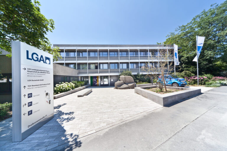 Das Bild zeigt ein modernes Bürogebäude mit drei Etagen und großen Fenstern, umgeben von Bäumen und Sträuchern. Im Vordergrund steht ein Schild mit Wegweisern sowie zwei Fahnen mit dem Logo „LGA“.