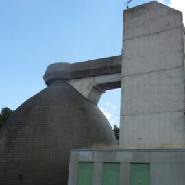Das Bild zeigt ein großes Industriegebäude mit einer markanten Kuppel aus dunklen Fliesen und einem angrenzenden hohen, rechteckigen Betonturm. Ein horizontaler Betonträger verbindet beide Bauwerke. Im Vordergrund befindet sich ein weiteres Gebäude mit grünen Türen und Fenstern, links im Hintergrund sind Bäume zu sehen.