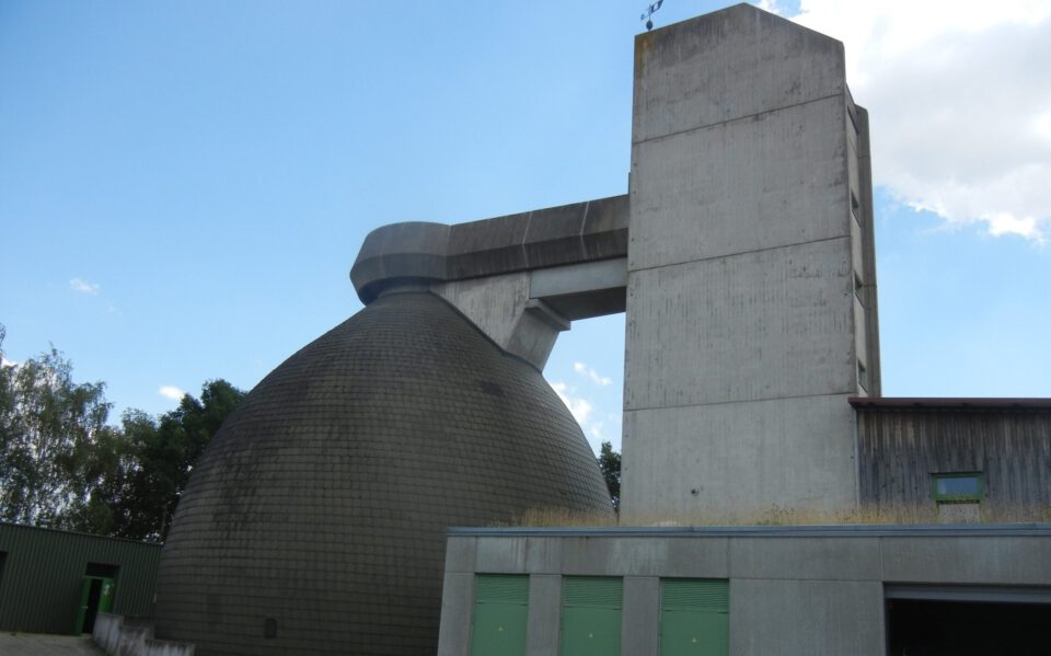 Das Bild zeigt ein großes Industriegebäude mit einer markanten Kuppel aus dunklen Fliesen und einem angrenzenden hohen, rechteckigen Betonturm. Ein horizontaler Betonträger verbindet beide Bauwerke. Im Vordergrund befindet sich ein weiteres Gebäude mit grünen Türen und Fenstern, links im Hintergrund sind Bäume zu sehen.