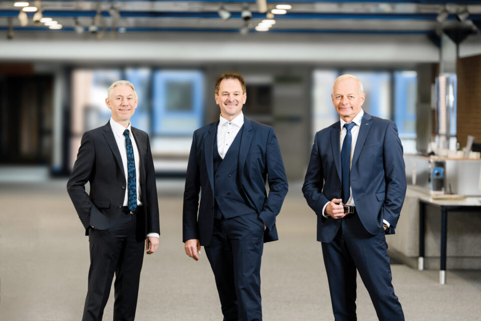 Das Bild zeigt drei Personen in einem modernen, großzügigen Innenraum. Alle tragen dunkle Anzüge mit weißen Hemden und Krawatten. Im Hintergrund sind eine hohe Decke mit sichtbaren Trägern, große Fenster sowie ein Tresen oder Empfangsbereich auf der rechten Seite zu sehen.