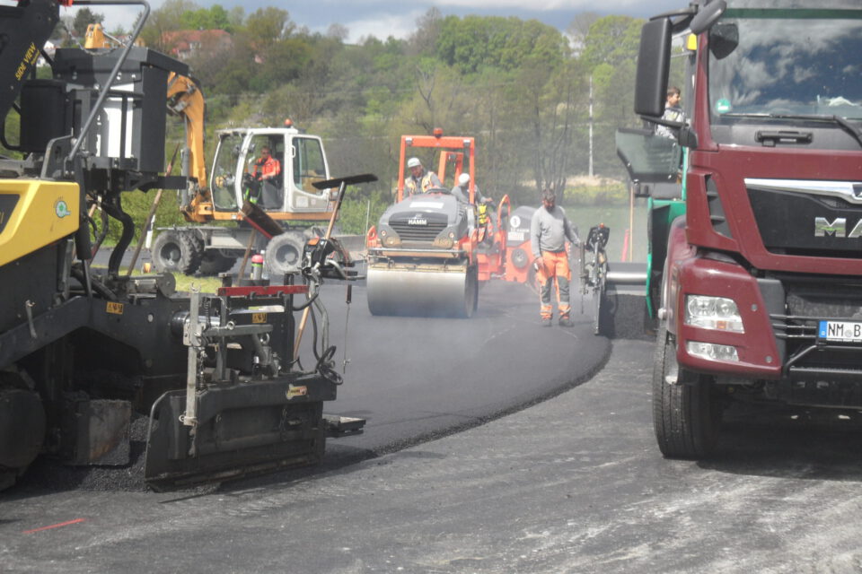 Das Bild zeigt eine Straßenszene mit mehreren Baustellenfahrzeugen und Arbeitern. Zu sehen sind ein gelber Asphaltfertiger, ein weißer Bagger im Hintergrund, eine orangefarbene Walze in der Mitte und ein roter Lkw auf der rechten Seite. Die Arbeiter sind mit dem Aufbringen von frischem Asphalt beschäftigt, im Hintergrund ist grüne Vegetation zu erkennen.