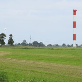 Das Bild zeigt eine ländliche Landschaft mit grünen Feldern im Vordergrund. Im Hintergrund sind mehrere Bäume, Windräder und ein hoher rot-weiß gestreifter Turm zu sehen. Der Himmel ist leicht bewölkt.