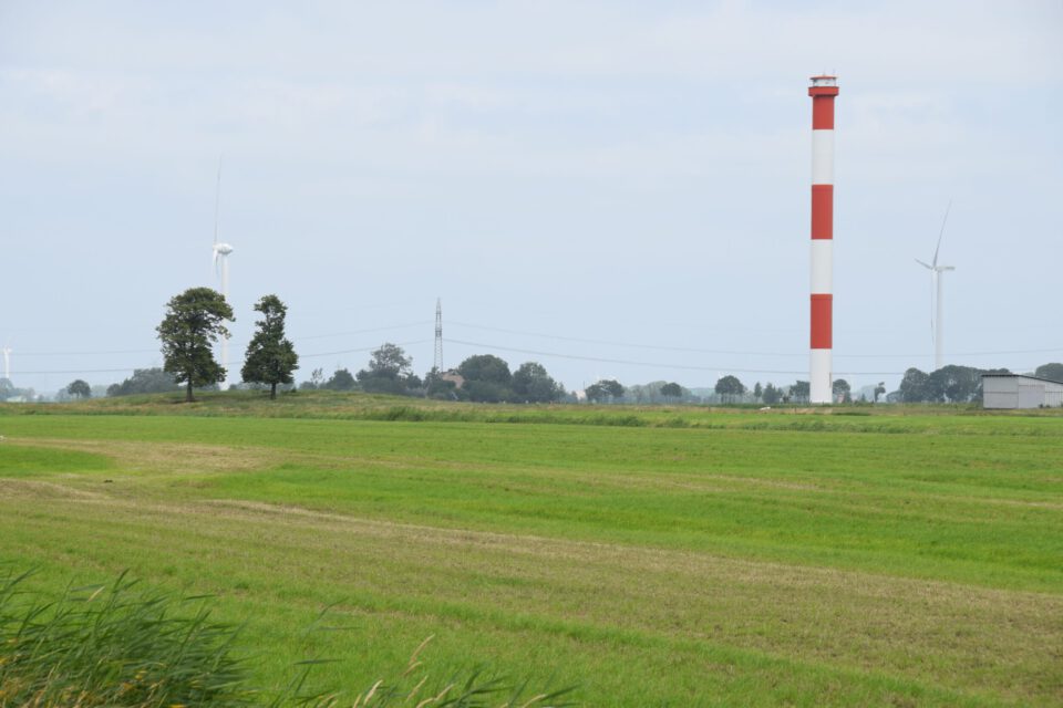 Das Bild zeigt eine ländliche Landschaft mit grünen Feldern im Vordergrund. Im Hintergrund sind mehrere Bäume, Windräder und ein hoher rot-weiß gestreifter Turm zu sehen. Der Himmel ist leicht bewölkt.
