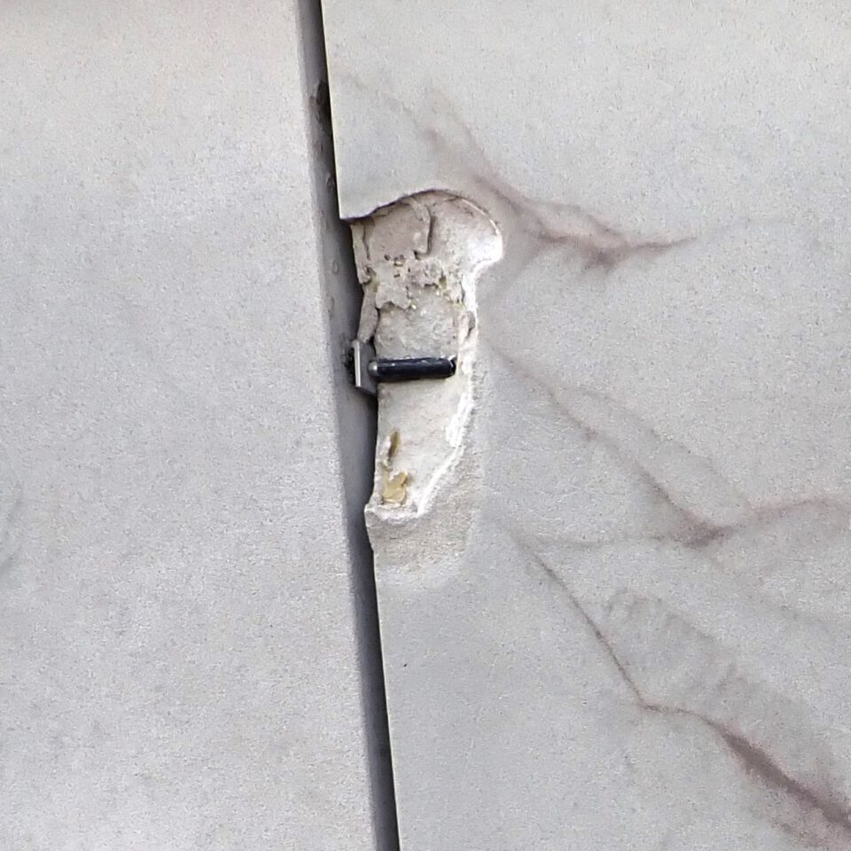 Das Bild zeigt eine Nahaufnahme einer beschädigten Wand- oder Paneelstelle mit einem sichtbaren Riss und Abplatzungen an der Verbindungsstelle zweier Platten. Ein Metallbeschlag oder Scharnier ist durch die Beschädigung teilweise freigelegt. Um das Bauteil herum sind Reste von Kleber oder Putz zu erkennen.