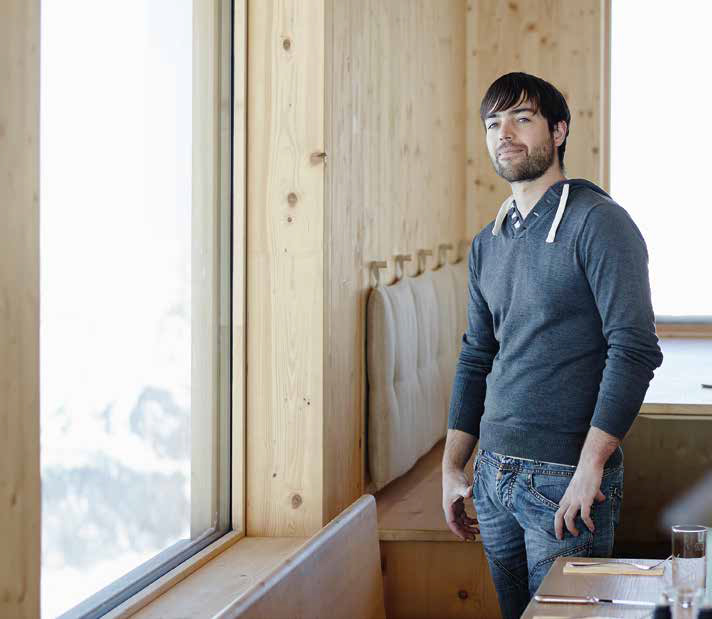 Das Bild zeigt eine Person in einem Raum mit Holzverkleidung, die aus einem großen Fenster blickt. Die Person trägt einen grauen Hoodie und Jeans. Entlang der Wand befinden sich gepolsterte Sitzgelegenheiten aus Holz.