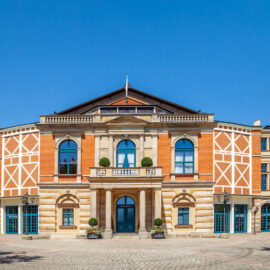 Das Bild zeigt ein großes, historisches Gebäude mit symmetrischer Fassade. In der Mitte befindet sich ein repräsentativer Eingang mit einem Rundbogen, flankiert von zwei Säulen und überdacht von einem Balkon. Das Gebäude besteht aus rotem Backstein und hellem Stein, mit weißen Verzierungen an den Seiten und mehreren Blumenkübeln vor dem Eingang.