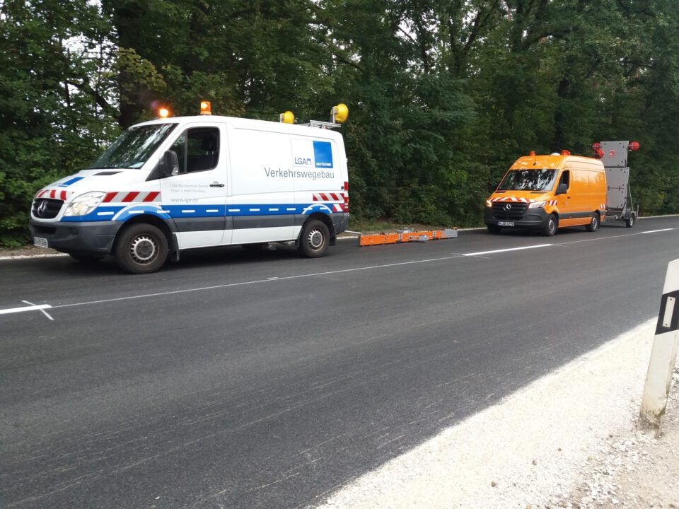 Das Bild zeigt zwei Fahrzeuge am Rand einer Straße. Das erste ist ein weißer Transporter mit blauen und roten Streifen, das zweite ein orangefarbener Transporter mit Anhänger.