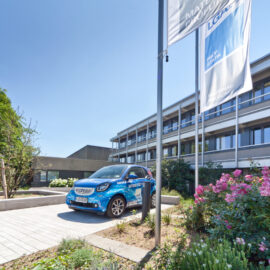 Das Bild zeigt die Außenansicht eines modernen mehrstöckigen Gebäudes mit großen Fenstern. Davor steht ein blauer Pkw auf einer gepflasterten Fläche, daneben befinden sich zwei hohe Fahnenmasten mit Fahnen. Die Umgebung ist mit Bäumen, Sträuchern und Blumen bepflanzt, der Himmel ist klar und blau.