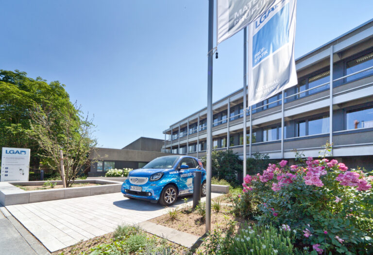 Das Bild zeigt die Außenansicht eines modernen mehrstöckigen Gebäudes mit großen Fenstern. Davor steht ein blauer Pkw auf einer gepflasterten Fläche, daneben befinden sich zwei hohe Fahnenmasten mit Fahnen. Die Umgebung ist mit Bäumen, Sträuchern und Blumen bepflanzt, der Himmel ist klar und blau.