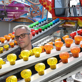 Das Bild zeigt eine Person, die hinter mehreren Reihen bunter Glühbirnen steht, die auf schrägen Metallschienen angeordnet sind. Die Glühbirnen leuchten in Farben wie Gelb, Orange, Rot und Grün. Im Hintergrund ist ein Teil eines Fahrgeschäfts mit einem lilafarbenen Wagen und weiteren bunten Dekorationen zu sehen.