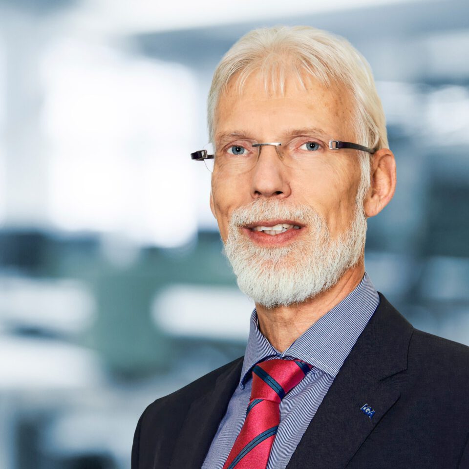 Das Bild zeigt eine Person, die eine Brille, ein dunkles Sakko, ein blaues Hemd mit feinen weißen Streifen und eine rot-blau gestreifte Krawatte trägt. Der Hintergrund ist unscharf und deutet auf eine Innenaufnahme, möglicherweise in einem Büro, hin.