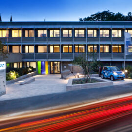 Das Bild zeigt ein modernes dreistöckiges Gebäude mit vielen beleuchteten Fenstern und flachem Dach, umgeben von Bäumen. Vor dem Gebäude befindet sich eine Einfahrt mit einem kleinen blauen Auto sowie ein Schild mit der Aufschrift „LGA“. Die Aufnahme entstand in der Dämmerung oder am frühen Abend.