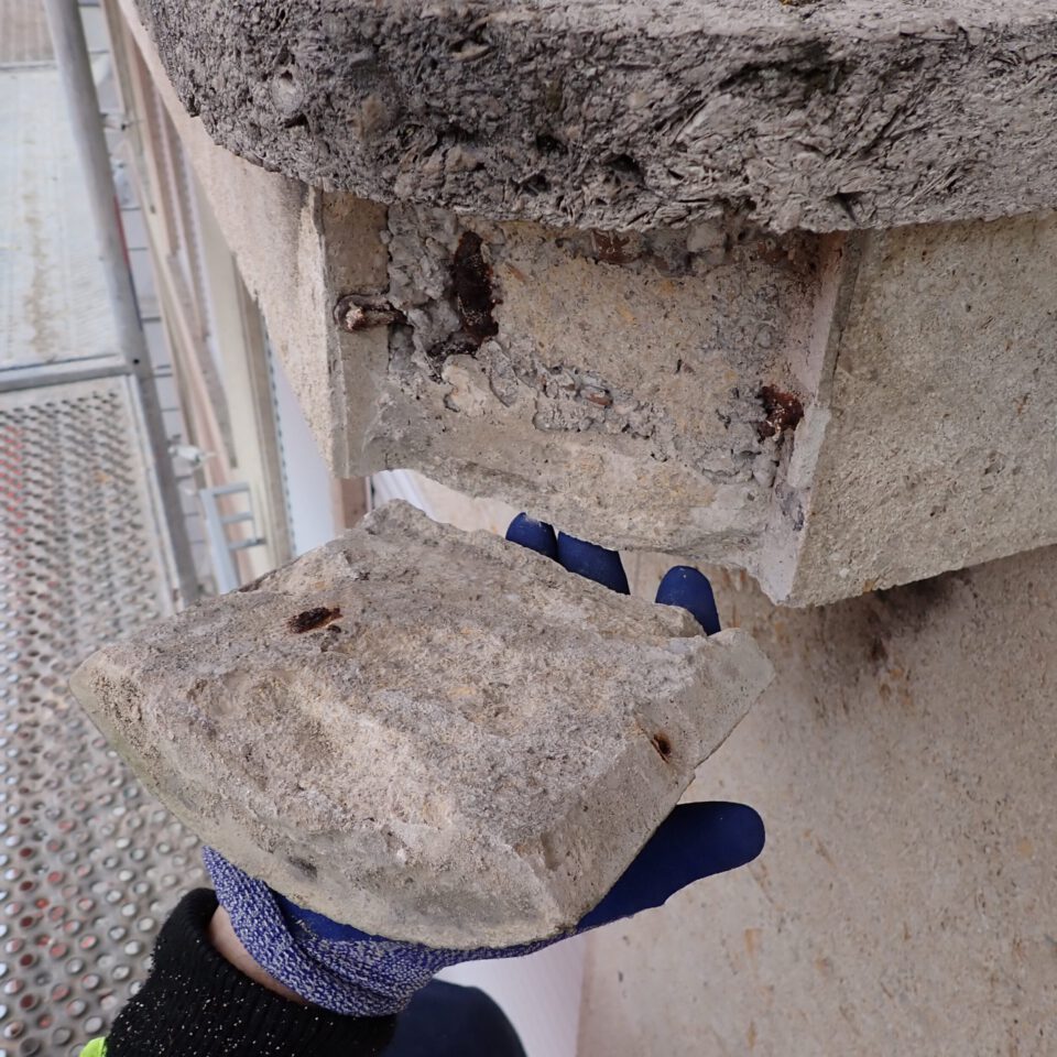 Das Bild zeigt eine Nahaufnahme einer behandschuhten Hand, die ein abgebrochenes Stück Beton hält. Im Inneren des Betonteils sind rostige Metallbewehrungen sichtbar. Im Hintergrund sind Teile eines Gebäudes und ein Gerüst zu erkennen.