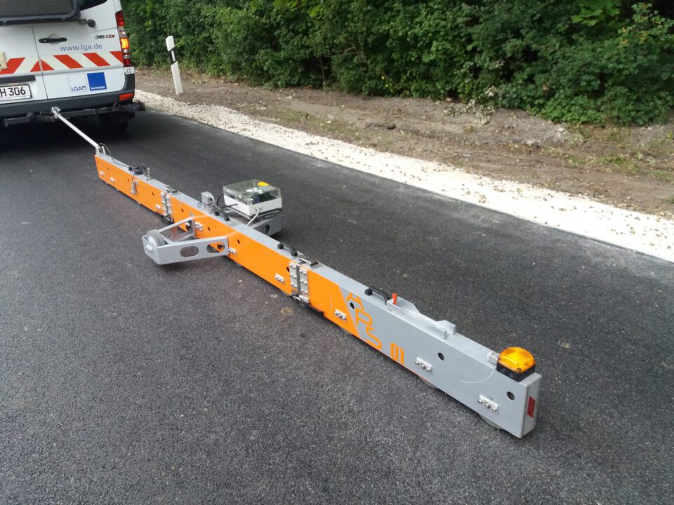 Das Bild zeigt ein Straßenvermessungsgerät, das am Heck eines Fahrzeugs angebracht ist und auf der Fahrbahn ausgefahren wurde. Es dient vermutlich der Messung oder Analyse des Straßenzustands. Das Fahrzeug trägt Markierungen und Hinweise auf eine Verbindung zu einem Unternehmen.