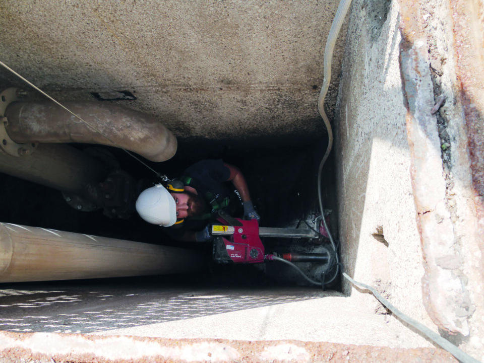 Das Bild zeigt eine Person mit weißem Schutzhelm und Sicherheitsausrüstung bei der Arbeit in einem engen Raum. Die Person bedient eine rote Bohrmaschine oder ein ähnliches Gerät. Umgeben ist sie von großen Rohren, Kabeln und Betonwänden.