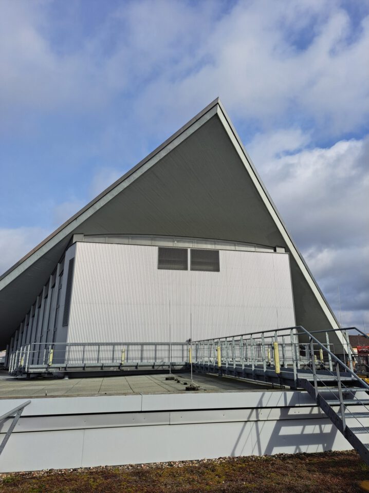 Das Bild zeigt ein großes, modernes Gebäude mit einem markanten, dreieckigen Dach. Die weiße Fassade ist von senkrechten Linien geprägt, und das Gebäude steht auf einer erhöhten Plattform mit Metallgeländern und Treppen. Darüber ist ein teils bewölkter Himmel zu sehen.