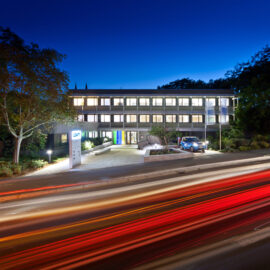 Das Bild zeigt ein modernes dreistöckiges Gebäude mit vielen beleuchteten Fenstern und flachem Dach, umgeben von Bäumen. Vor dem Gebäude befindet sich eine Einfahrt mit einem kleinen blauen Auto sowie ein Schild mit der Aufschrift „LGA“. Die Aufnahme entstand in der Dämmerung oder am frühen Abend.