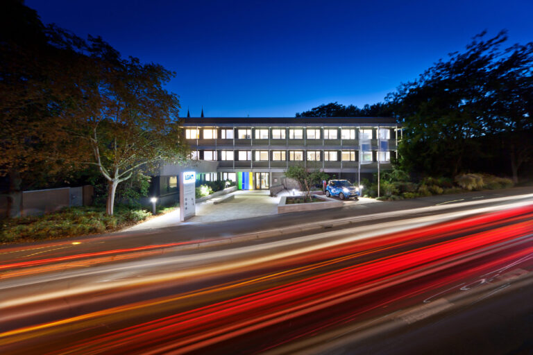 Das Bild zeigt ein modernes dreistöckiges Gebäude mit vielen beleuchteten Fenstern und flachem Dach, umgeben von Bäumen. Vor dem Gebäude befindet sich eine Einfahrt mit einem kleinen blauen Auto sowie ein Schild mit der Aufschrift „LGA“. Die Aufnahme entstand in der Dämmerung oder am frühen Abend.