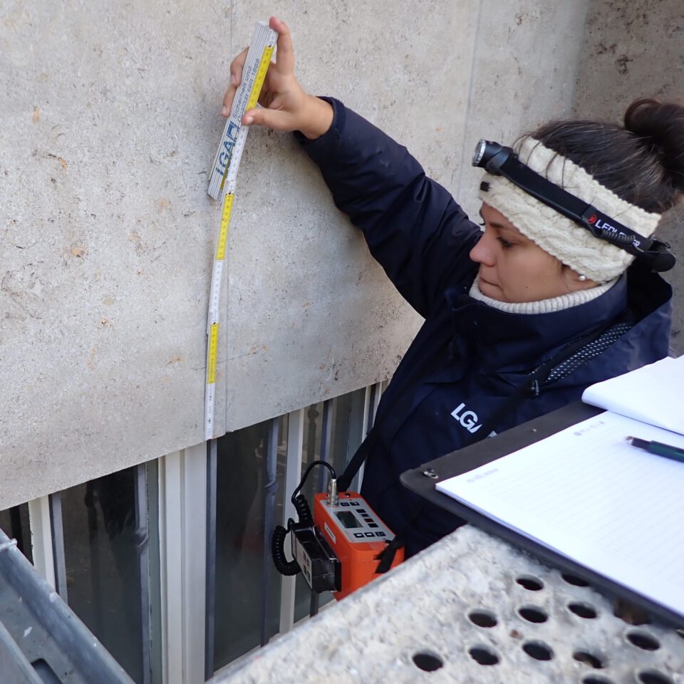 Das Bild zeigt eine Person mit Stirnlampe, die eine Messlatte an die Außenwand eines Gebäudes hält. Die Person steht auf einem Gerüst oder einer Plattform, im Vordergrund liegt ein Klemmbrett mit Papieren und einem Stift. Das Gesicht der Person ist aus Datenschutzgründen unkenntlich gemacht.