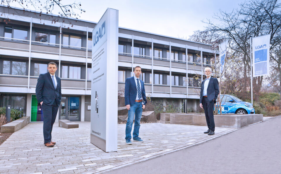 Das Bild zeigt drei Personen in Businesskleidung vor einem Gebäude mit der Aufschrift „LGA“. Im Hintergrund steht ein Auto mit dem LGA-Logo, daneben befinden sich Fahnen mit dem gleichen Logo.