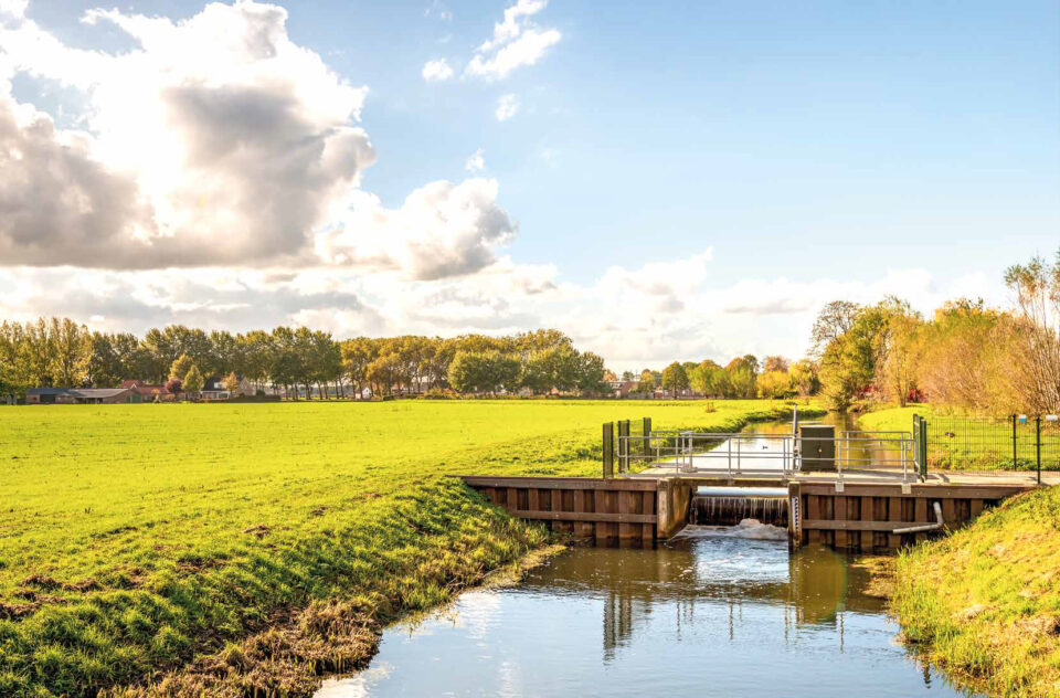 Das Bild zeigt eine Landschaft mit einem wasserbaulichen Bauwerk im Vordergrund, vermutlich ein Wehr oder eine Schleusenanlage. Im Hintergrund befinden sich Wiesen und Bäume unter einem teils bewölkten Himmel.