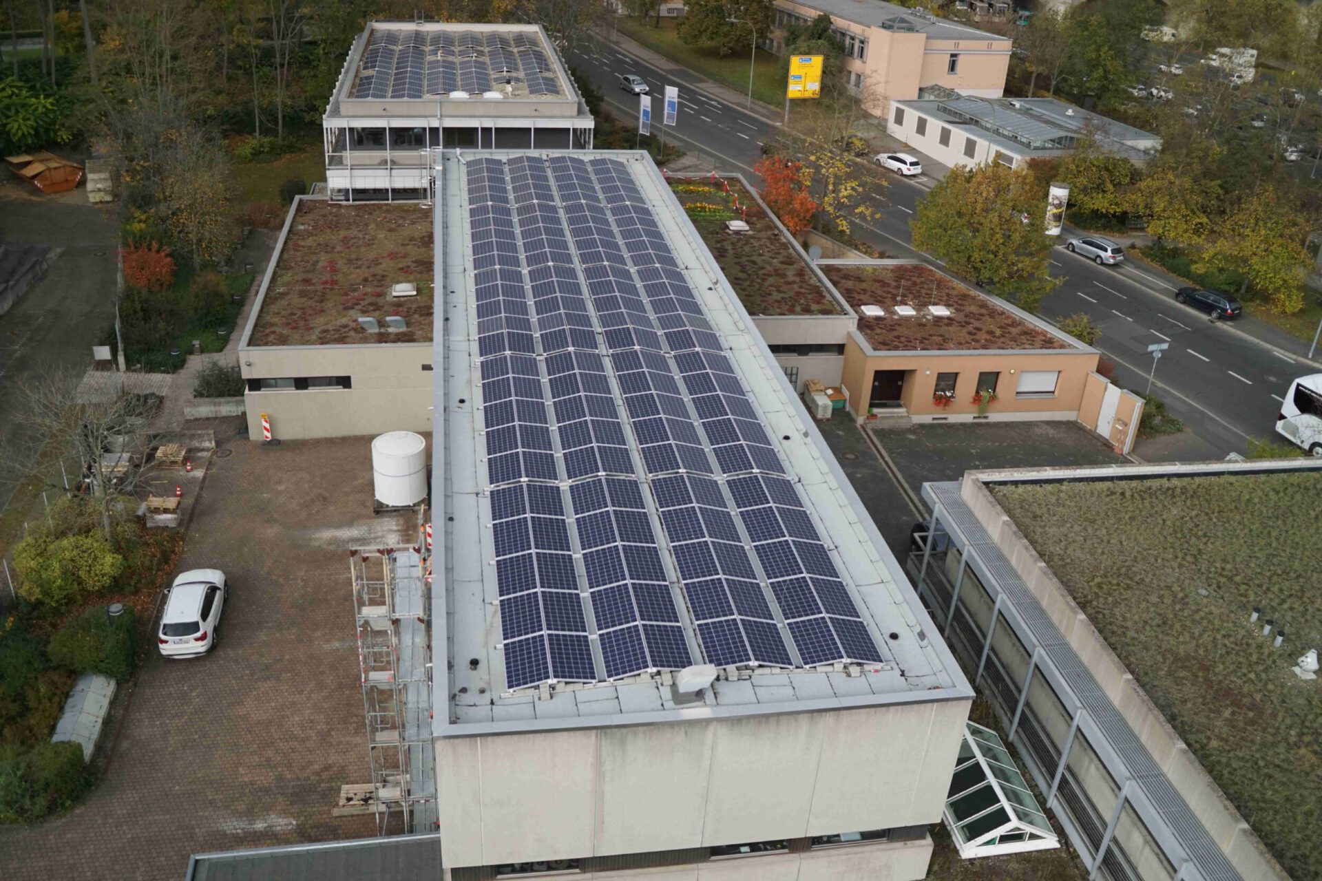 Das Bild zeigt eine Luftaufnahme eines Gebäudekomplexes mit mehreren Flachdachbauten. Im Zentrum befindet sich ein großes Gebäude mit einer umfangreichen Solaranlage auf dem Dach. Einige umliegende Gebäude haben begrünte Dächer, teils ebenfalls mit Solarpaneelen, und unten links ist ein Parkplatz mit einem weißen Auto zu sehen.