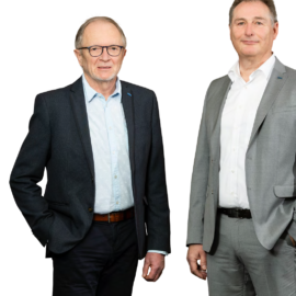 Das Bild zeigt zwei Personen in Businesskleidung, die nebeneinander stehen. Die linke Person trägt ein dunkles Sakko, ein hellblaues Hemd und eine dunkle Hose mit braunem Gürtel. Die rechte Person trägt einen hellgrauen Anzug mit weißem Hemd und schwarzem Gürtel.