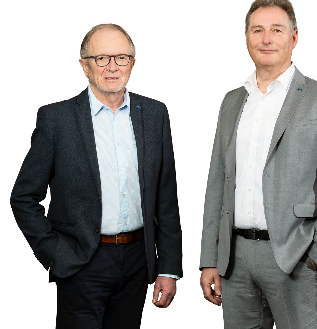 Das Bild zeigt zwei Personen in Businesskleidung, die nebeneinander stehen. Die linke Person trägt ein dunkles Sakko, ein hellblaues Hemd und eine dunkle Hose mit braunem Gürtel. Die rechte Person trägt einen hellgrauen Anzug mit weißem Hemd und schwarzem Gürtel.