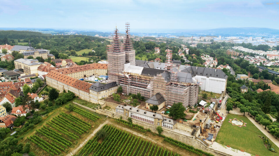 Das Bild zeigt eine Luftaufnahme eines großen historischen Gebäudekomplexes, der sich in Renovierung befindet. Mehrere Türme und Gebäudeteile sind eingerüstet, im Vordergrund sind Weinberge zu sehen, rechts ein Parkbereich. Im Hintergrund erstreckt sich eine Stadtlandschaft mit Wohnhäusern und weiteren Gebäuden.