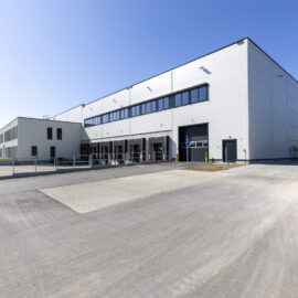 Das Bild zeigt ein großes Industrie- oder Lagergebäude mit moderner weißer Fassade, mehreren Laderampen und großen Fenstern im oberen Bereich. Vor dem Gebäude befindet sich eine weitläufige asphaltierte Fläche für Fahrzeuge. Der Himmel ist klar und blau.