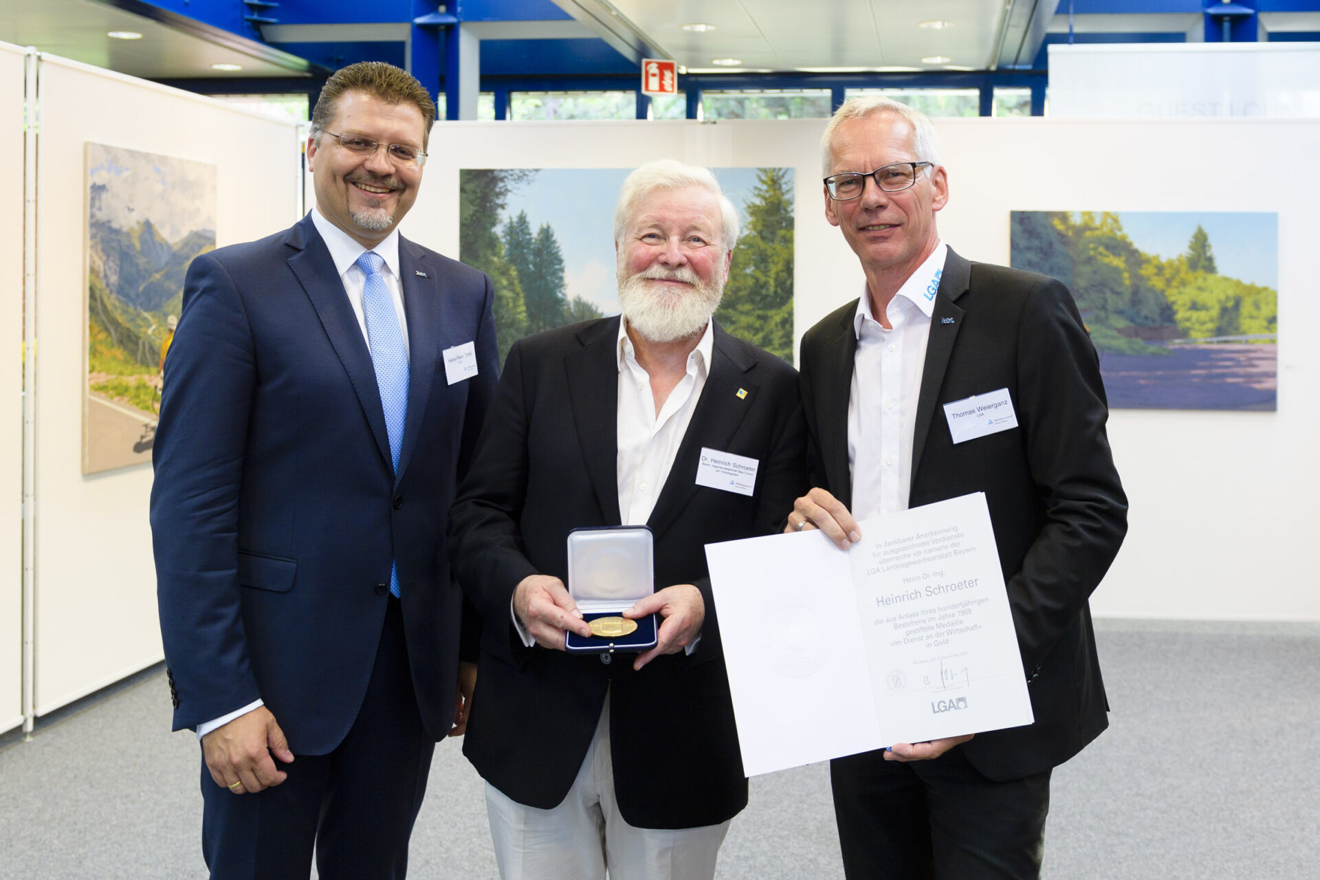 Das Bild zeigt drei Männer in Anzügen, die in einem formellen Rahmen zusammenstehen. Der Mann in der Mitte hält eine geöffnete Schachtel mit einer Medaille, der Mann rechts ein Zertifikat. Im Hintergrund sind zwei Landschaftsgemälde und weiße Trennwände zu sehen.