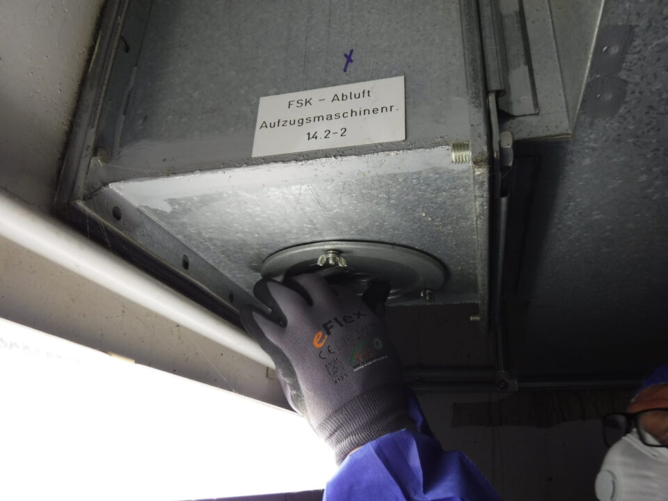 Eine Person mit Schutzhandschuhen und entsprechender Kleidung arbeitet an einem Gerät, das an der Decke montiert ist. Es handelt sich um eine technische Anlage mit sichtbaren Bauteilen und einem Hinweisetikett.