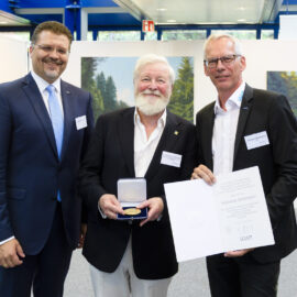 Das Bild zeigt drei Männer in Anzügen, die in einem formellen Rahmen zusammenstehen. Der Mann in der Mitte hält eine geöffnete Schachtel mit einer Medaille, der Mann rechts ein Zertifikat. Im Hintergrund sind zwei Landschaftsgemälde und weiße Trennwände zu sehen.