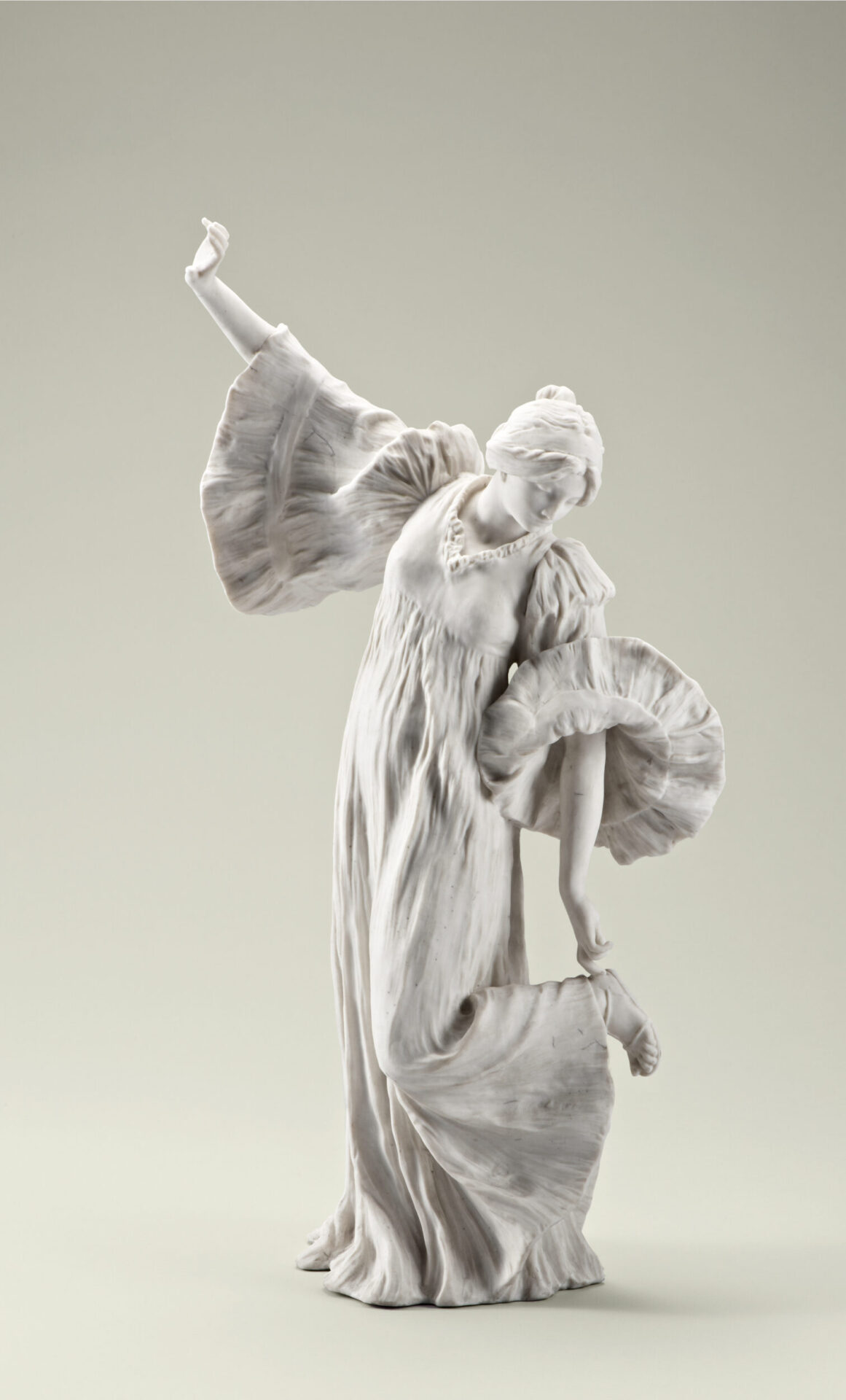 Das Bild zeigt eine weiße Skulptur aus Marmor oder Porzellan, die eine Person in einem fließenden Kleid darstellt. Die Figur hält mit einem Arm einen Teil des Kleides, während der andere Arm erhoben ist, und der Kopf ist leicht nach unten geneigt. Das Kleid weist detaillierte Falten und Rüschen auf, besonders an den Ärmeln und am Saum.