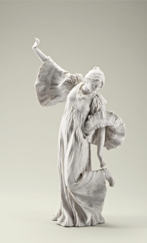 Das Bild zeigt eine weiße Skulptur aus Marmor oder Porzellan, die eine Person in einem fließenden Kleid darstellt. Die Figur hält mit einem Arm einen Teil des Kleides, während der andere Arm erhoben ist, und der Kopf ist leicht nach unten geneigt. Das Kleid weist detaillierte Falten und Rüschen auf, besonders an den Ärmeln und am Saum.