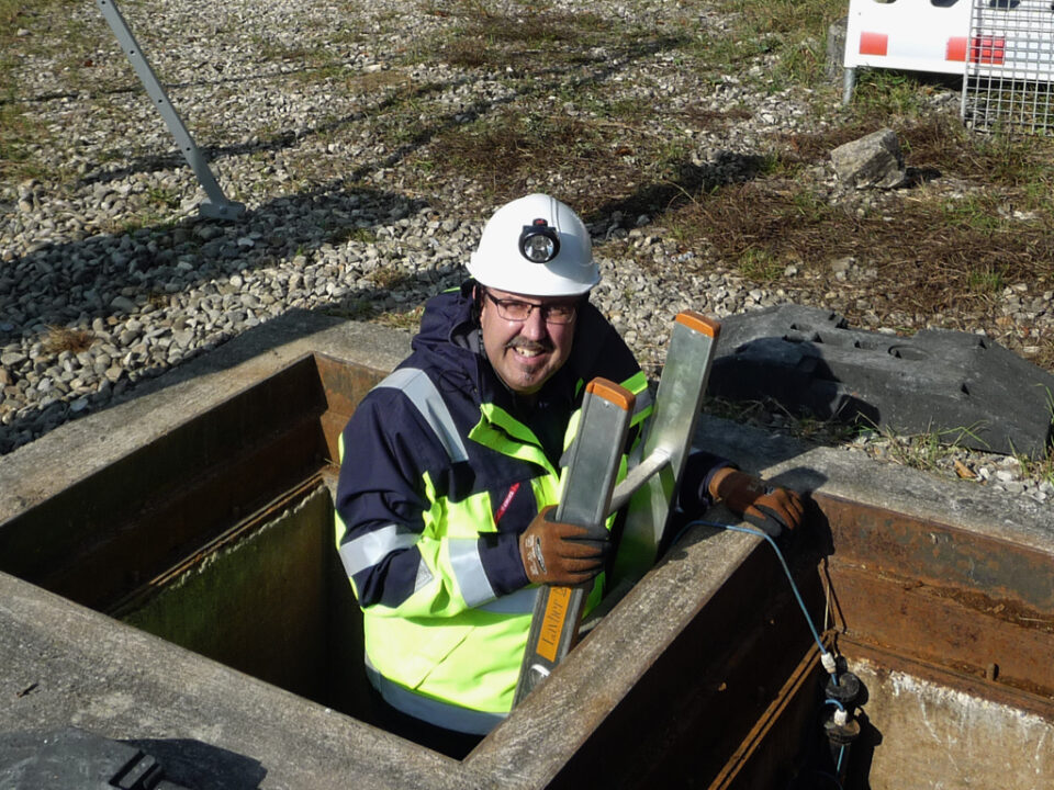 Eine Person mit Warnjacke und weißem Schutzhelm mit Stirnlampe steigt aus einem unterirdischen Versorgungsschacht. Sie hält sich an einer Metallleiter fest, im Hintergrund sind Kies, etwas Vegetation und ein Teil einer Baustellenabsperrung zu sehen.