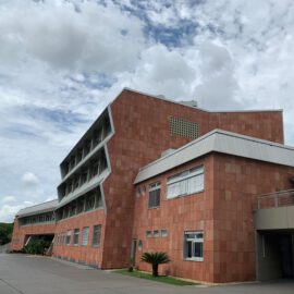 Das Bild zeigt ein großes, modern gestaltetes Gebäude mit mehreren Etagen und einer Fassade aus rötlich-braunen Fliesen. Die Fenster sind teilweise mit weißen Klappläden versehen, das Dach ist schräg und hat graue Kanten. Der Himmel ist teils bewölkt, am Fuß des Gebäudes ist eine kleine Pflanze zu sehen.
