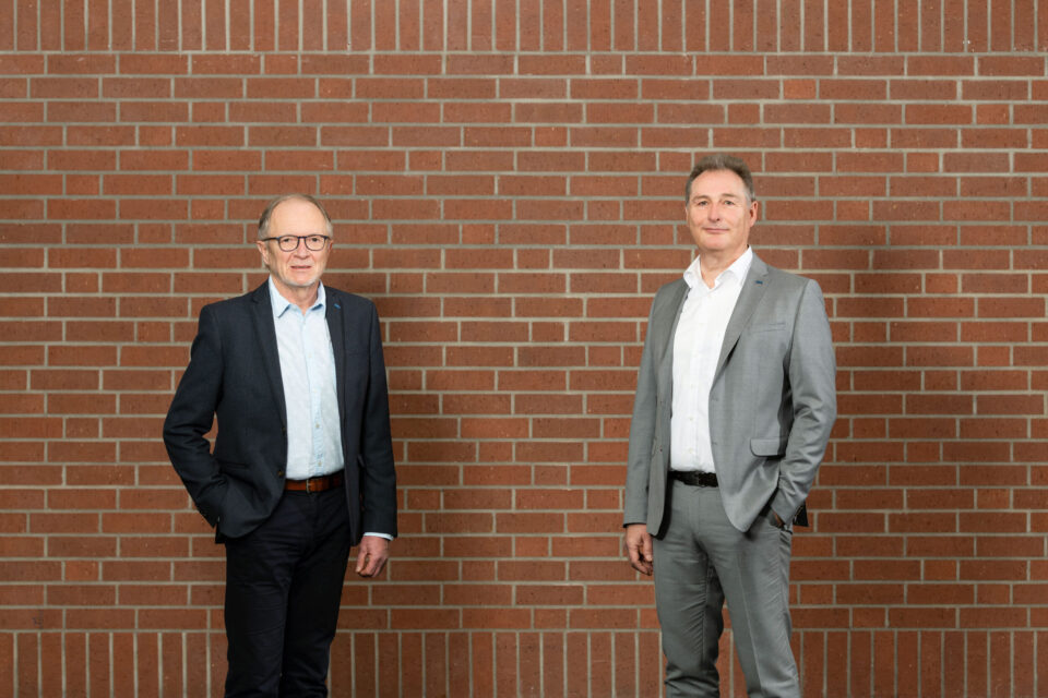 Das Bild zeigt zwei Personen in formeller Kleidung, die vor einer Ziegelwand stehen. Die linke Person trägt einen dunklen Anzug mit hellblauem Hemd, die rechte einen hellgrauen Anzug mit weißem Hemd.