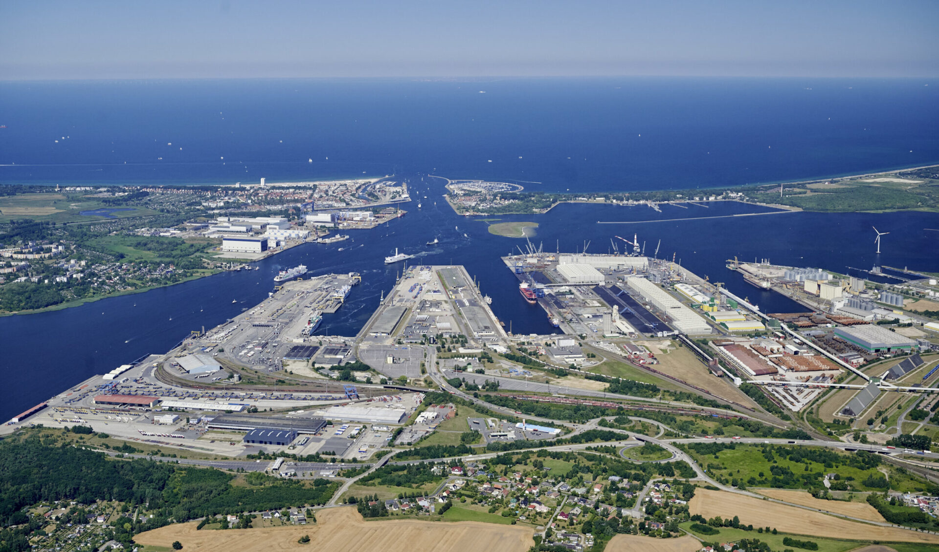 Das Bild zeigt eine Luftaufnahme eines großen Hafengebiets mit mehreren Anlegestellen, Lagerhallen und Industriegebäuden entlang eines Flusses, der ins Meer mündet. Mehrere Schiffe liegen an den Kais, und Kräne sind in den Industrieabschnitten zu erkennen. Die Umgebung umfasst städtische Bereiche, Grünflächen und landwirtschaftlich genutzte Felder, im Hintergrund ist eine weite Wasserfläche mit vielen kleinen Booten zu sehen.