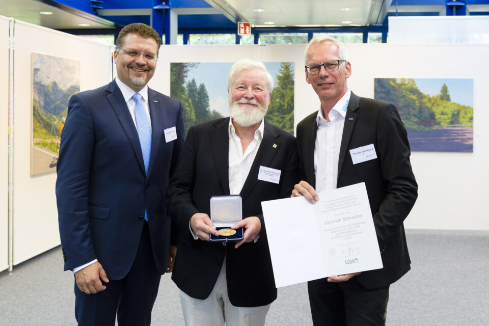 Das Bild zeigt drei Männer in Anzügen, die in einem formellen Rahmen zusammenstehen. Der Mann in der Mitte hält eine geöffnete Schachtel mit einer Medaille, der Mann rechts ein Zertifikat. Im Hintergrund sind zwei Landschaftsgemälde und weiße Trennwände zu sehen.