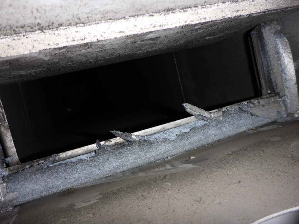 Das Bild zeigt eine Nahaufnahme des Inneren eines Lüftungskanals oder Schachts. Die metallische Oberfläche ist mit Staub und Schmutz bedeckt, an den Rändern sind beschädigte oder abgebrochene Stellen zu erkennen. Das Innere des Kanals ist dunkel und nur schwer einsehbar.