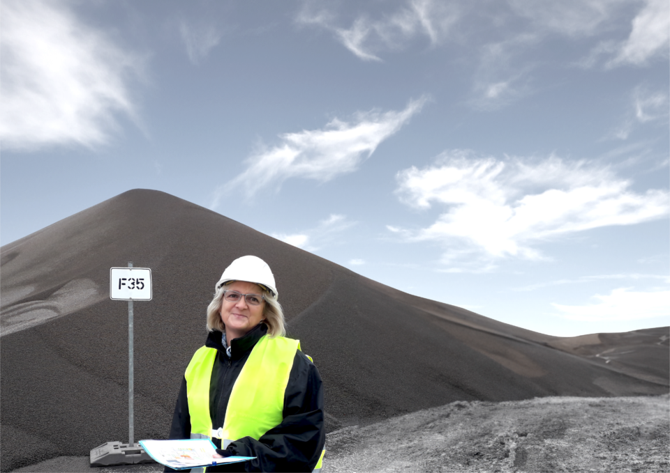Das Bild zeigt eine Person mit weißem Schutzhelm und gelber Warnweste, die vor einem großen Kies- oder Schotterhaufen steht. Die Person hält Unterlagen oder Pläne in der Hand. Im Hintergrund ist ein teils bewölkter Himmel zu sehen.