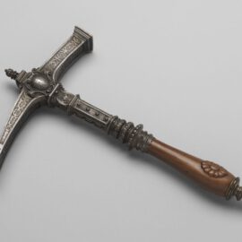 Das Bild zeigt einen kunstvoll verzierten Kriegshammer mit einem Metallkopf und einem hölzernen Griff. Der Hammerkopf ist mit filigranen Gravuren und floralen Mustern geschmückt. Der Griff ist glatt gearbeitet, mit geschnitzten Details versehen und endet in einem abgerundeten Knauf mit weiteren Verzierungen.