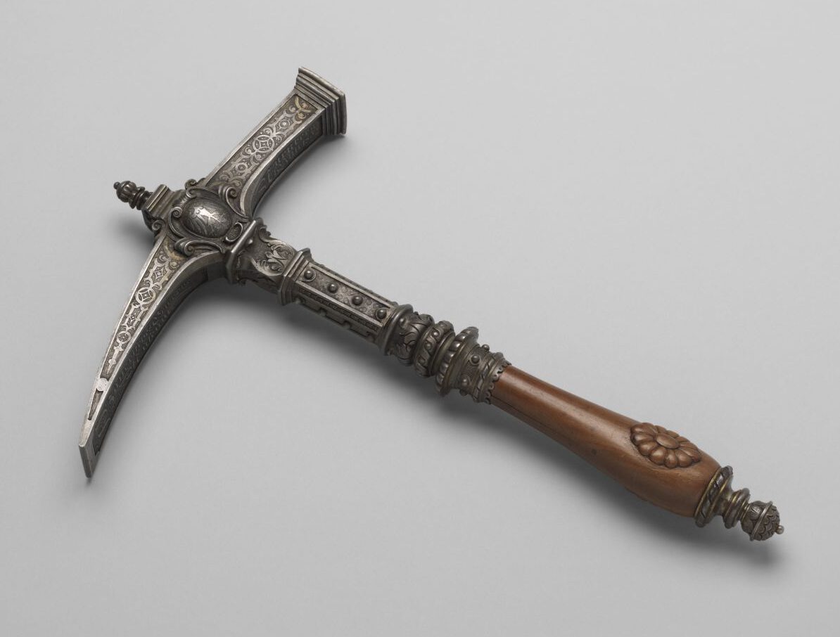 Das Bild zeigt einen kunstvoll verzierten Kriegshammer mit einem Metallkopf und einem hölzernen Griff. Der Hammerkopf ist mit filigranen Gravuren und floralen Mustern geschmückt. Der Griff ist glatt gearbeitet, mit geschnitzten Details versehen und endet in einem abgerundeten Knauf mit weiteren Verzierungen.