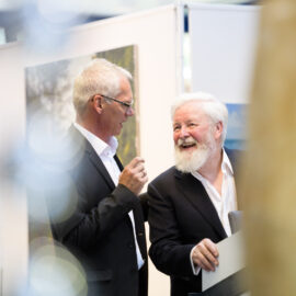 Das Bild zeigt zwei Personen in Anzügen, die miteinander im Gespräch sind. Im Hintergrund sind Ausstellungstafeln und Kunstwerke zu sehen.